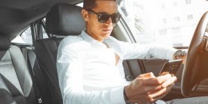 Man with sunglasses looking at phone in one hand while holding steering wheel from other